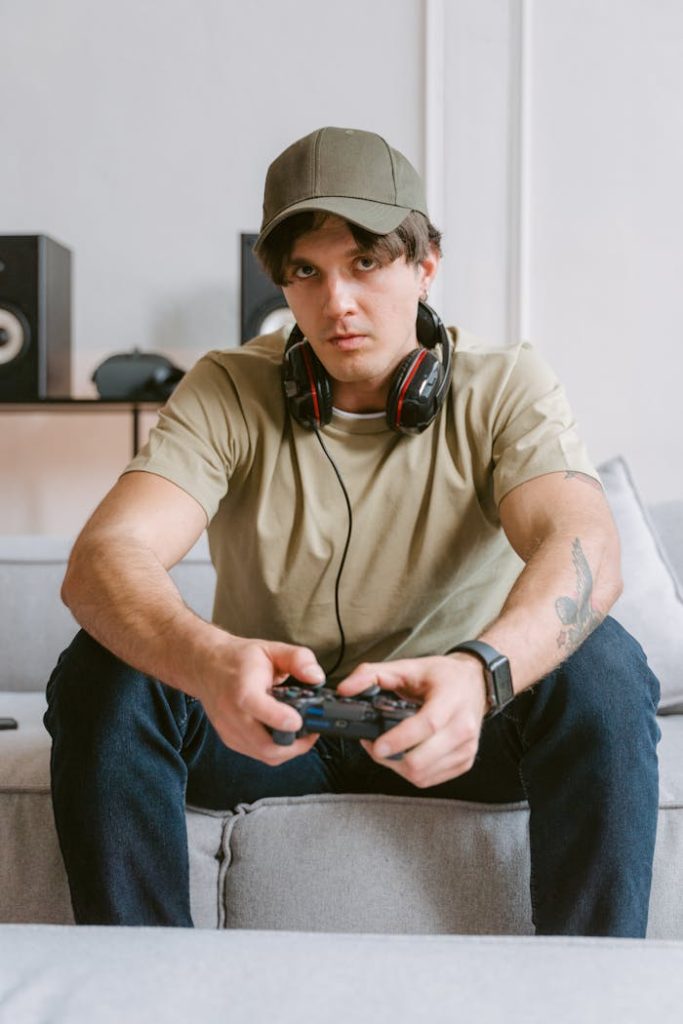 Casual young man gaming on a couch wearing headphones and a cap indoors.